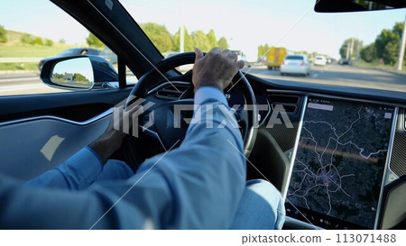 Hands of businessman holds steering wheel while driving electric car at city. Male businessperson operating electrical vehicle on urban road. Young man commuting to work at sunny day. Close up 113071488