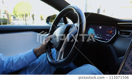 Hands of businessman holds steering wheel while driving electric car at city. Male businessperson operating electrical vehicle on urban road. Young man commuting to work at sunny day. Close up Hands of businessman holds steering wheel while driving electric car at city. Male businessperson operating electrical vehicle on urban road. Young man commuting to work at sunny day. Close up 113071506