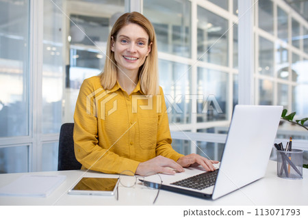 Confident female professional typing on a laptop in a modern office, feeling happy and empowered at work. 113071793