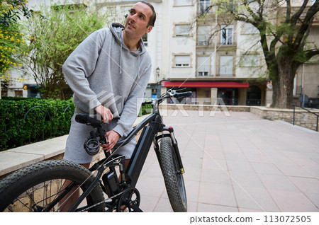 Young sporty man adjusts the height of new battery powered bike, electric bicycle before taking it out for first ride 113072505