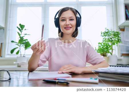 Webcam portrait of young female student wearing headphones, sitting at home at desk 113074420