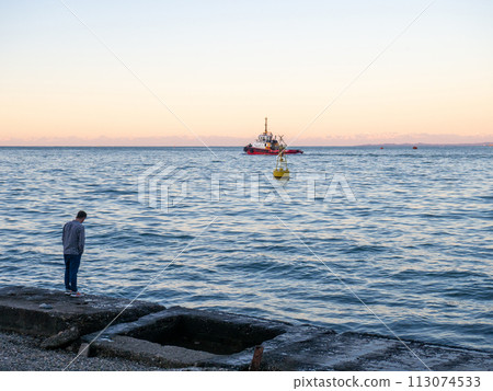 Port area. Tugboat on the road. A buoy in the bay of the port. Marine urban landscape. A man stands on a concrete pier Port area. Tugboat on the road. A buoy in the bay of the port. Marine urban landscape. A man stands on a concrete pier 113074533