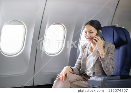 Young asian woman sitting on seat in airplane and talking by smartphone. Young woman traveller talking on cell phone Young asian woman sitting on seat in airplane and talking by smartphone. Young woman traveller talking on cell phone 113074807