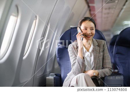 Young asian woman sitting on seat in airplane and talking by smartphone. Young woman traveller talking on cell phone Young asian woman sitting on seat in airplane and talking by smartphone. Young woman traveller talking on cell phone 113074808