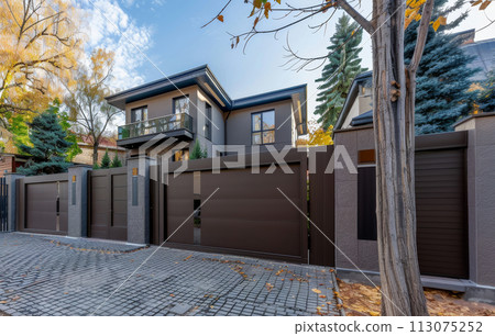 A gray private house of two floors hidden behind a fence with automatic sliding brown gates A gray private house of two floors hidden behind a fence with automatic sliding brown gates 113075252