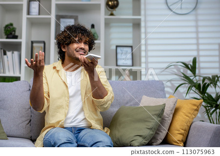 A young man in a casual outfit sits on the couch at home, talking on his mobile phone with a puzzled look, surrounded by cozy cushions and indoor plants. A young man in a casual outfit sits on the couch at home, talking on his mobile phone with a puzzled look, surrounded by cozy cushions and indoor plants. 113075963