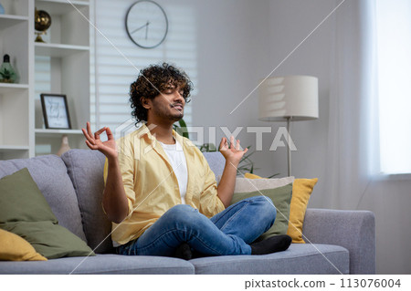 Peaceful Indian man with curly hair meditating in lotus pose on a sofa at home, exhibiting calmness and relaxation. 113076004