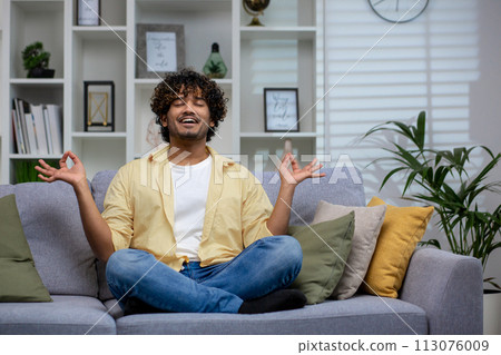 A young adult sits cross-legged on a couch, meditating in a peaceful living room setting, embodying tranquility and mindfulness. A young adult sits cross-legged on a couch, meditating in a peaceful living room setting, embodying tranquility and mindfulness. 113076009