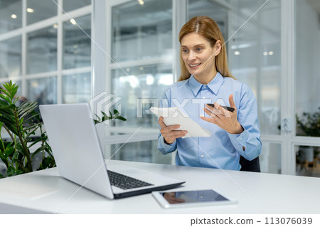 A woman in a modern office space is focused on learning and taking notes while working on her digital tablet. A woman in a modern office space is focused on learning and taking notes while working on her digital tablet. 113076039