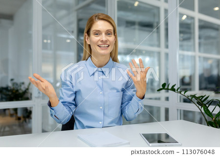 A professional woman in a blue shirt gestures animatedly while looking at a camera, implying a video call discussion. A professional woman in a blue shirt gestures animatedly while looking at a camera, implying a video call discussion. 113076048