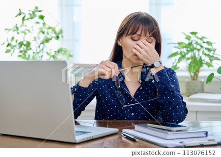 Tired sad mature woman at workplace at table with computer laptop Tired sad mature woman at workplace at table with computer laptop 113076152