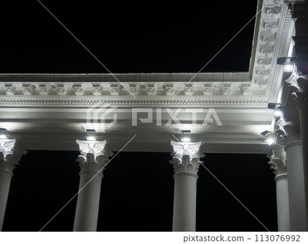 White colonnade in the dark. White columns in the light of spotlights. Street decorative architecture. Element of the architectural ensemble in the city park. White colonnade in the dark. White columns in the light of spotlights. Street decorative architecture. Element of the architectural ensemble in the city park. 113076992