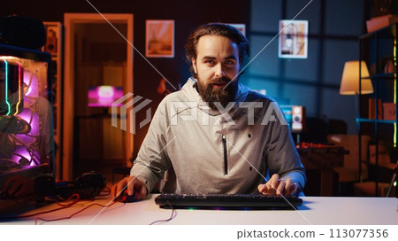 Man turning gaming chair around, starting livestream, greeting audience, talking about strategies to beat online opponents. Smiling gamer streaming live from RGB lit living room, chatting with fans 113077356