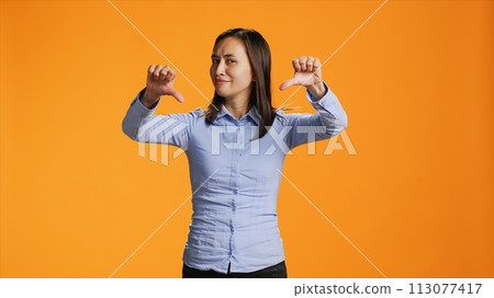 Asian person giving thumbs down in front of studio camera, expressing negativity and disapproval with dislike symbol. Negative woman showing rejection, being upset and displeased. 113077417