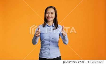 Asian model presenting thumbs up symbol in studio, expressing good vibes and success against orange background. Young smiling person giving like sign on camera, shows approval gesture. 113077420