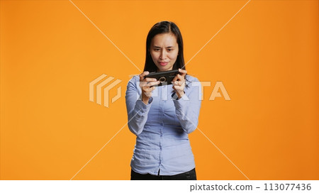 Joyful adult playing mobile videogames on camera, having fun with modern shooting competition online on smartphone network. Asian woman using phone for entertainment and leisure. Joyful adult playing mobile videogames on camera, having fun with modern shooting competition online on smartphone network. Asian woman using phone for entertainment and leisure. 113077436