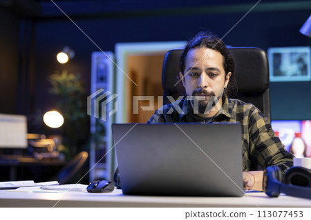 Young man typing email report on his laptop, researching and preparing presentations. Male university student working with paperwork research, browsing online website and finishing assignments. Young man typing email report on his laptop, researching and preparing presentations. Male university student working with paperwork research, browsing online website and finishing assignments. 113077453