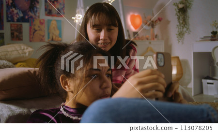 Asian teen lies on bed and watches content with friend 113078205