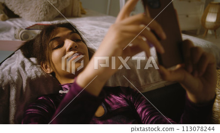Girl sits near bed, smiles and surfs internet using phone 113078244