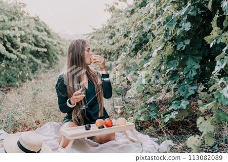 Woman picnic vineyard. Happy woman with a glass of wine at a picnic in the vineyard, wine tasting at sunset and open nature in the summer. Romantic dinner, fruit and wine. 113082089