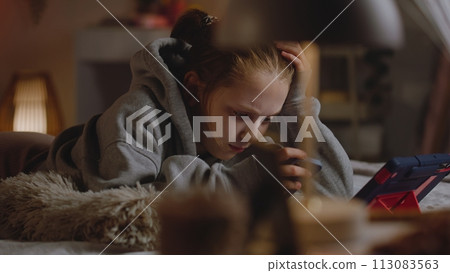 Young girl lies on the bed in cozy bedroom, chats online using phone Young girl lies on the bed in cozy bedroom, chats online using phone 113083563