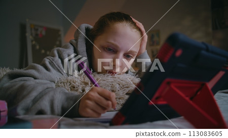 Girl writes in notebook and does online school lessons at home using digital tablet Girl writes in notebook and does online school lessons at home using digital tablet 113083605