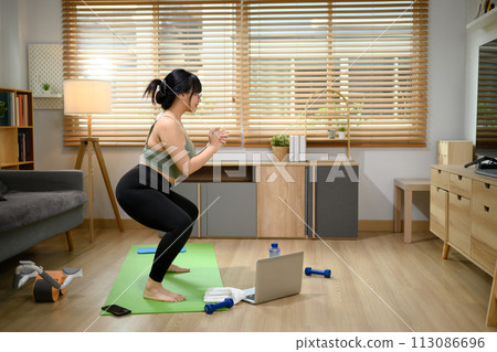 Portrait of young woman doing squats on exercise mat during home workout Portrait of young woman doing squats on exercise mat during home workout 113086696