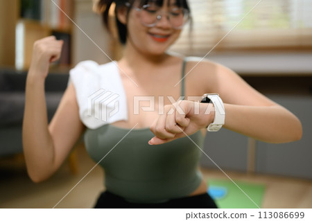 Smiling young woman checking results on smartwatch after home workout Smiling young woman checking results on smartwatch after home workout 113086699