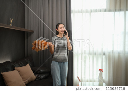 Pretty young housewife with dust brush talking on mobile phone in living room 113086716