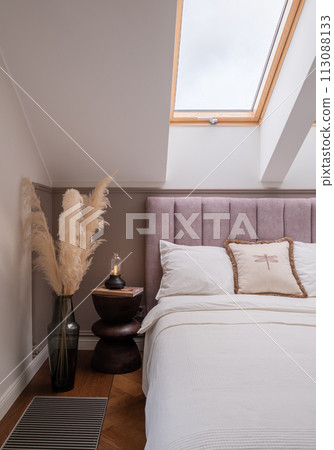 A bedroom with a bed, nightstand, vase of pampas grass, and a skylight fixture 113088133