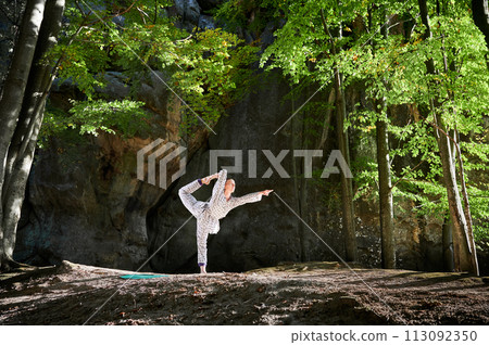 Woman practicing yoga outdoors in forest. Barefoot female on yoga mat surrounded by trees and large rocks, which suggests peaceful, natural environment ideal for meditation or yoga practice. 113092350
