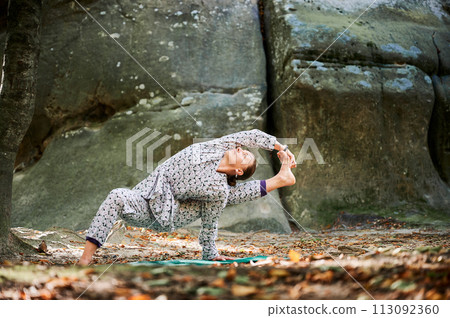 Woman practicing yoga outdoors in forest. Barefoot female on yoga mat surrounded by trees and large rocks, which suggests peaceful, natural environment ideal for meditation or yoga practice. 113092360