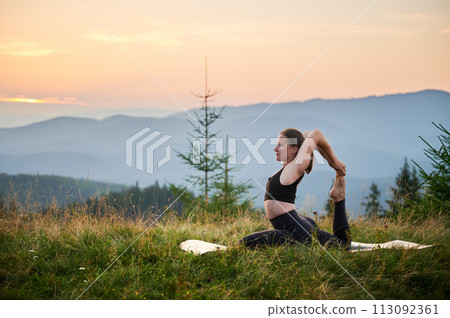 Woman practicing yoga outdoors in the mountains in a serene, natural setting. Female performing yoga pose on mat, with backdrop of beautiful mountain landscape at sunrise or sunset. 113092361