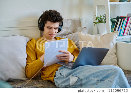 Young guy college student in headphones using laptop sitting on couch at home Young guy college student in headphones using laptop sitting on couch at home 113096713