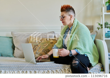 Mature woman with blanket holding cup looking at laptop sitting on couch at home 113096716