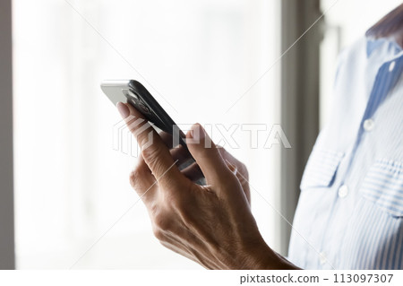 Mature female hands holding black smartphone against window 113097307