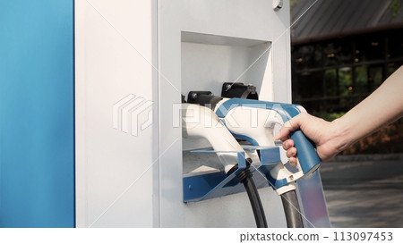 Hand holding EV charging plug in front of camera with blurred background of outdoor. Driver of electric car hold batteries charger plug. Sustainable energy powered electric charging station background Hand holding EV charging plug in front of camera with blurred background of outdoor. Driver of electric car hold batteries charger plug. Sustainable energy powered electric charging station background 113097453