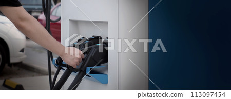 Hand holding EV charging plug in front of camera with blurred background of outdoor. Driver of electric car hold batteries charger plug. Sustainable energy powered electric charging station background 113097454