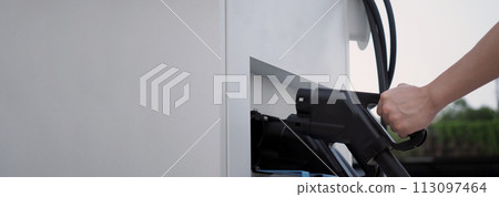 Hand holding EV charging plug in front of camera with blurred background of outdoor. Driver of electric car hold batteries charger plug. Sustainable energy powered electric charging station background Hand holding EV charging plug in front of camera with blurred background of outdoor. Driver of electric car hold batteries charger plug. Sustainable energy powered electric charging station background 113097464