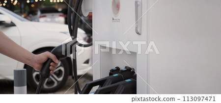 Hand holding EV charging plug in front of camera with blurred background of outdoor. Driver of electric car hold batteries charger plug. Sustainable energy powered electric charging station background 113097471