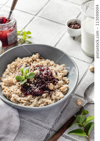 Oatmeal porridge with raspberry jam and nuts in gray bowl close up on gray tile background served with spoon. Healthy diet breakfast 113099004