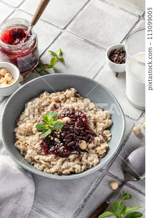 Oatmeal porridge with raspberry jam and nuts in gray bowl on gray tile background served with spoon. Healthy diet breakfast 113099005