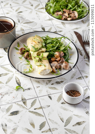 French rolled omelette stuffed with cream cheese with avocado, aragula and mushrooms for breakfast on white plate printed tile background, text space 113099011