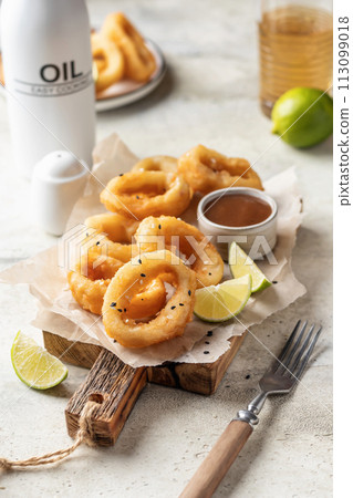Crisp fried golden squid rings with lemon and sauce on wooden board, light background 113099018
