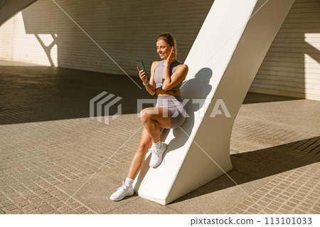 A young woman in sportswear is resting after a workout outside while standing with smartphone 113101033
