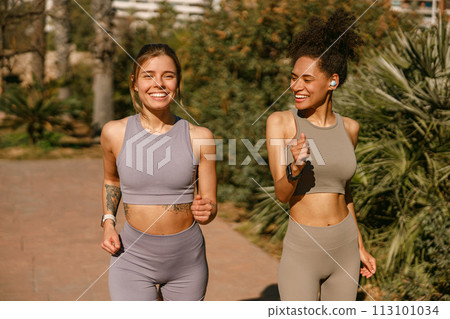 Two active women athlete running side by side along an outdoor track on public park background Two active women athlete running side by side along an outdoor track on public park background 113101034