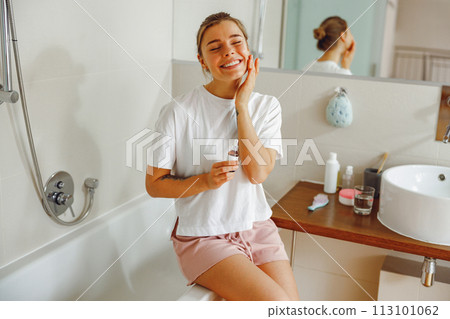 Beautiful young woman applying cosmetic serum onto her face in bathroom. Facial Skincare 113101062