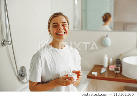 Young smiling woman holding jar with skincare cream in hands. Home beauty routine Young smiling woman holding jar with skincare cream in hands. Home beauty routine 113101100