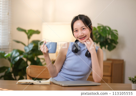 Young attractive happy Asian female student holding a cup of coffee sitting in the living room smiling and looking at the camera while working on a laptop at the home office. Young attractive happy Asian female student holding a cup of coffee sitting in the living room smiling and looking at the camera while working on a laptop at the home office. 113105555