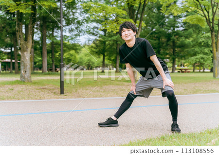 A man in sportswear bends and stretches his knees during warm-up before exercising in a park A man in sportswear bends and stretches his knees during warm-up before exercising in a park 113108556